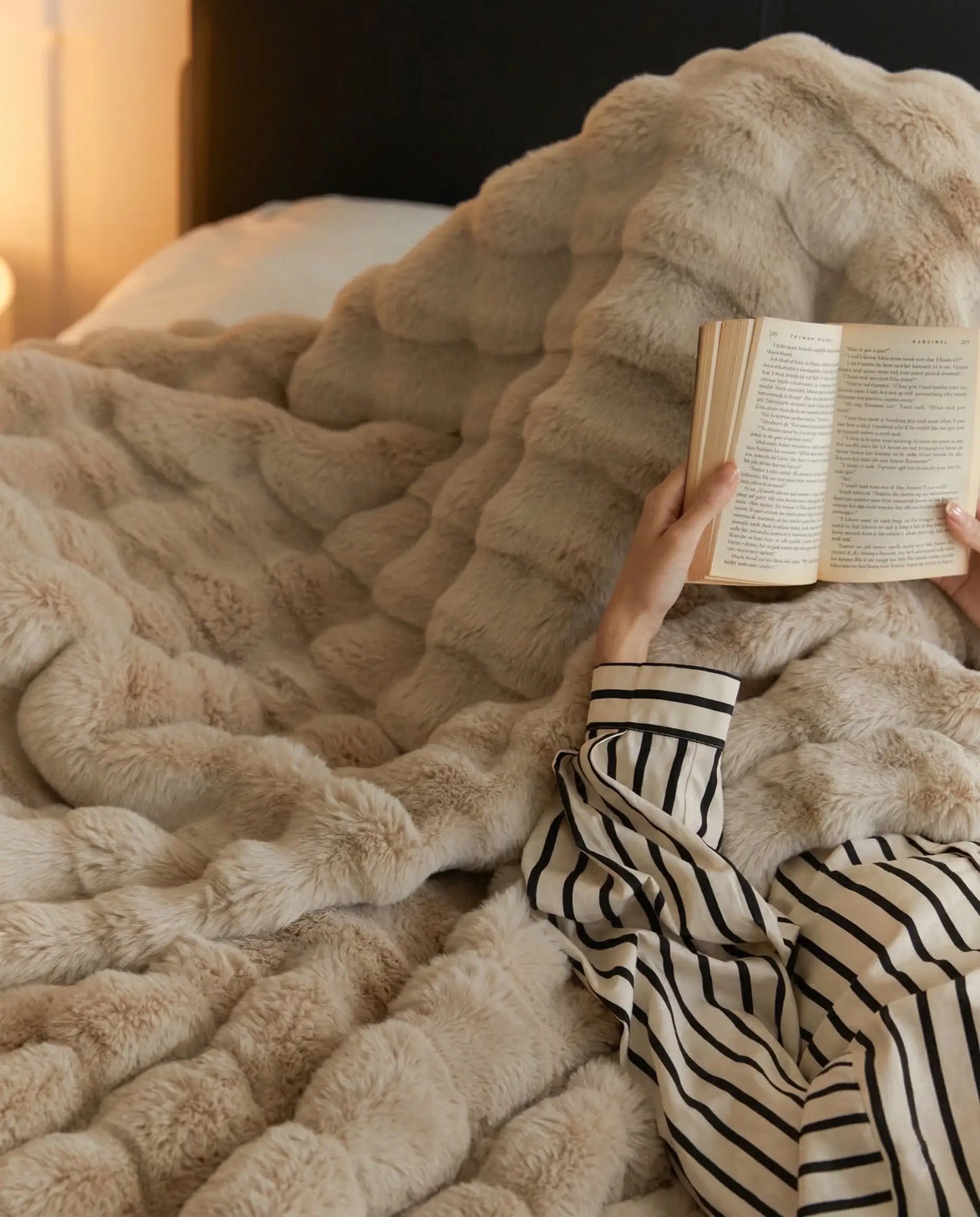 Person reading book in bed wrapped in DORMÉ Studio Marshmallow Ultra Plush Blanket khaki plush texture.