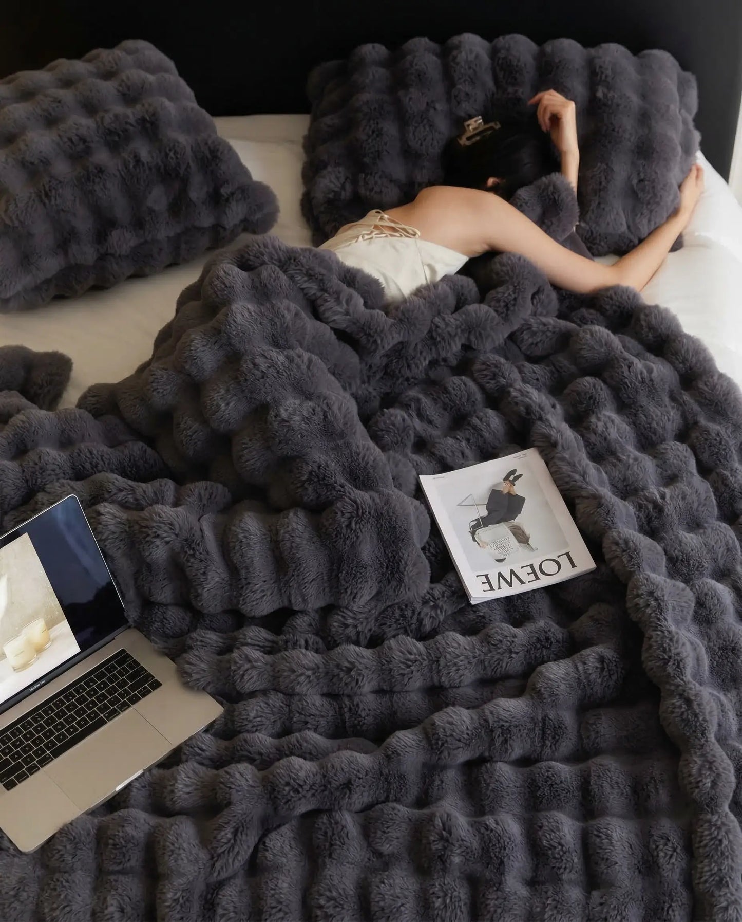 Woman resting in bed under DORMÉ Studio Marshmallow Ultra Plush Blanket charcoal plush texture with laptop and magazine.