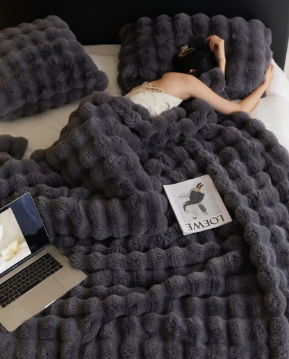 Woman resting in bed under DORMÉ Studio Marshmallow Ultra Plush Blanket charcoal plush texture with laptop and magazine.