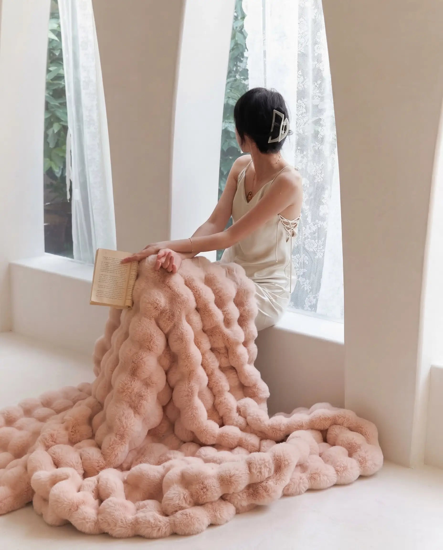 Woman sitting by window wrapped in DORMÉ Studio Marshmallow Ultra Plush Blanket pink plush fabric reading a book.